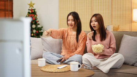 Photo for Young woman lesbian couple in sweater watching cinema with excited face and holding tv remote control to switching cinema channel while eating delicious popcorn and spending time together. - Royalty Free Image