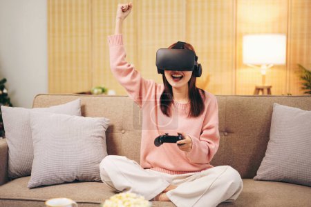 Foto de Mujer joven lesbiana usando suéter y gafas VR mientras sostiene el joystick para jugar a videojuegos con experiencia en realidad virtual y levantando el brazo para celebrar la victoria mientras se relaja en la sala de estar. - Imagen libre de derechos