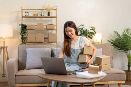 Photo for Young entrepreneur is using laptop to checking online orders and holding cardboard boxes while sitting on comfortable the couch and working in living room at home office. - Royalty Free Image