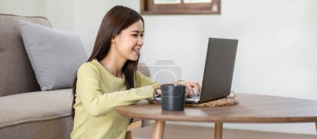 Téléchargez les photos : Jeune femme est assis à sourire sur le sol et en utilisant un ordinateur portable pour dactylographier rapport d'entreprise tout en travaillant et en magasinant en ligne pour se détendre dans le salon à la maison. - en image libre de droit