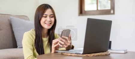 Téléchargez les photos : Jeune femme est assis à sourire sur le sol et en utilisant un ordinateur portable pour dactylographier rapport d'entreprise tout en travaillant et en se relaxant avec des achats en ligne sur smartphone dans le salon à la maison. - en image libre de droit