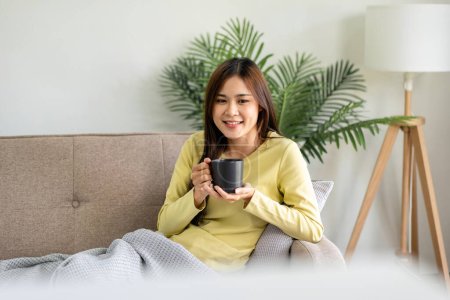 Foto de Joven mujer asiática está bebiendo café y tumbado en el gran sofá cómodo para ver películas en la televisión en la sala de estar en casa. - Imagen libre de derechos