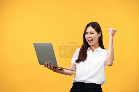 Téléchargez les photos : Jeune femme asiatique portant une chemise blanche à manches courtes et tenant un ordinateur portable pour travailler tout en souriant et en levant un bras pour célébrer après avoir réussi à travailler isolé sur fond jaune. - en image libre de droit