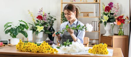Téléchargez les photos : Fleuriste féminine en tablier utilisant des ciseaux pour préparer et couper la tige de chrysanthème blanc pour la création et la fabrication de bouquets de fleurs dans sa boutique de fleurs. - en image libre de droit