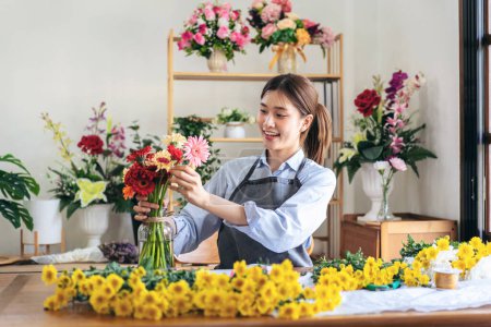 Photo for Female florist in apron smiling and arranging gerbera flowers in glass vase with enjoying while creating and designing floral bouquet in her flower shop. - Royalty Free Image