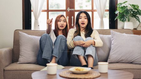 Foto de Mujer joven pareja lesbiana viendo películas de terror y señalando con la cara emocionada mientras pasan tiempo para relajarse juntos en el gran sofá cómodo en casa. - Imagen libre de derechos