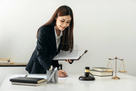 Photo for Business lawyer woman holding business contract to reading and writing data on document while working on the table with brass scales and justice hammer in law firm. - Royalty Free Image