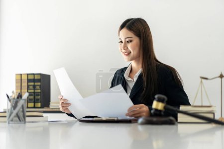 Photo for Business lawyer woman holding business contract to reading while sitting to working on the table with brass scales and justice hammer in law firm office. - Royalty Free Image
