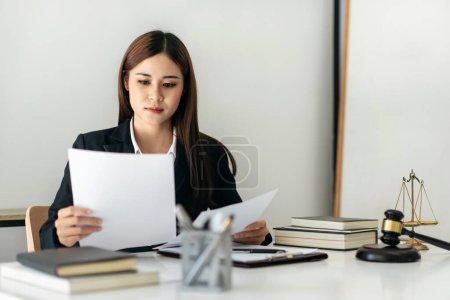 Photo for Business lawyer woman holding business contract to reading while sitting to working on the table with brass scales and justice hammer in law firm office. - Royalty Free Image