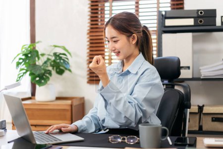 Photo for Businesswoman is reading about accounting of business with good news and raising fist with delightful while using laptop to working and typing information of new business project in modern office. - Royalty Free Image