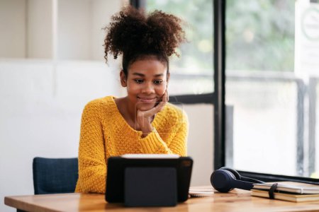 Téléchargez les photos : Femme étudiante afro-américaine en pull utilisant une tablette numérique pour regarder l'apprentissage en ligne et étudier la leçon en ligne tout en étant assise sur le bureau pour apprendre les connaissances et l'éducation à l'université. - en image libre de droit