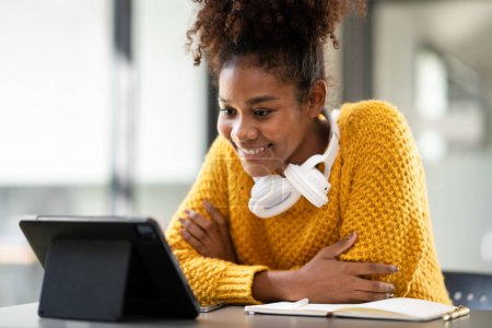 Téléchargez les photos : Étudiante afro-américaine en pull regardant l'apprentissage en ligne pour étudier la leçon en ligne sur tablette et prendre des notes tout en étant assise sur le bureau pour apprendre les connaissances et l'éducation à l'université. - en image libre de droit