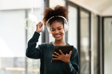 Téléchargez les photos : Étudiante afro-américaine portant un pull et un casque pour étudier la leçon en ligne et écrire des informations sur une tablette numérique tout en apprenant l'éducation et les connaissances à l'université. - en image libre de droit