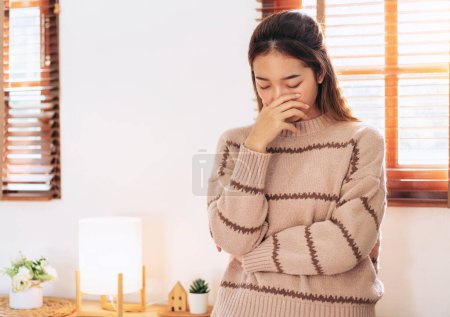 Foto de Mujer en suéter que cubre la nariz con la mano mientras se siente enfermo y dolor de cabeza con migraña durante el descanso y de pie en la sala de estar en casa. - Imagen libre de derechos