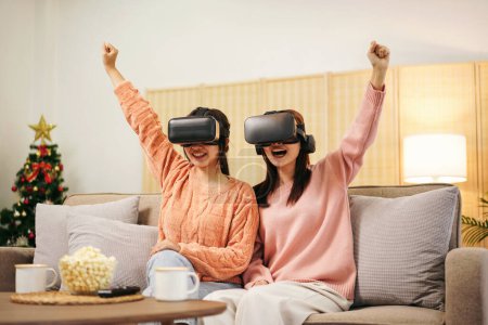 Foto de Mujer joven pareja lesbiana que usa suéter y gafas VR para levantar el brazo con la cara emocionada mientras ve la experiencia de la realidad virtual con disfrutar y pasar tiempo juntos en la sala de estar en casa. - Imagen libre de derechos