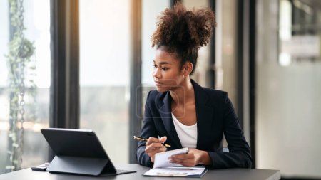 African american businesswoman in suit use digital tablet to reading accounting information of business and taking notes in notebook while working and thinking strategy of project in modern workspace. Poster 646177432
