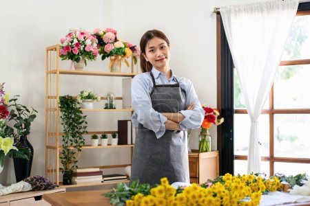 Photo for Female florist in apron smiling and standing with arms crossed in her flower shop to creating and designing floral for arrangement flower bouquet with chrysanthemum on the table. - Royalty Free Image