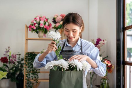 Téléchargez les photos : Fleuriste féminine en tablier arrangeant chrysanthème blanc dans un sac en papier tout en créant et en concevant floral pour arrangement bouquet de fleurs dans sa boutique de fleurs. - en image libre de droit