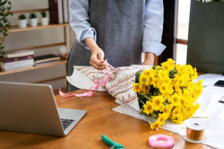 Photo for Female florist in apron using craft paper to wrapping yellow chrysanthemum and tie with pink ribbon on the table while creating and designing floral for arrangement flower bouquet in her flower shop. - Royalty Free Image