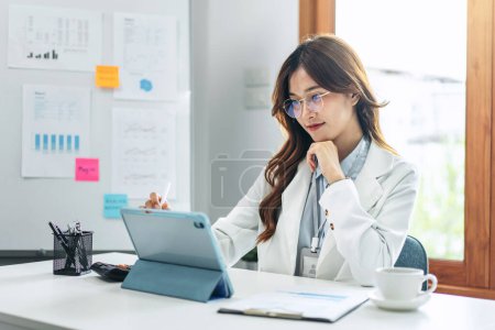 Photo for Businesswoman in suit is reading business data for thinking and analysis about strategy and planning of new business while working and writing on tablet of technology device in modern office. - Royalty Free Image