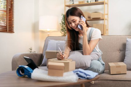 Photo for Young entrepreneur is talking with customer on smartphone and writing address on cardboard boxes while sitting on comfortable the couch and working in living room at home office. - Royalty Free Image