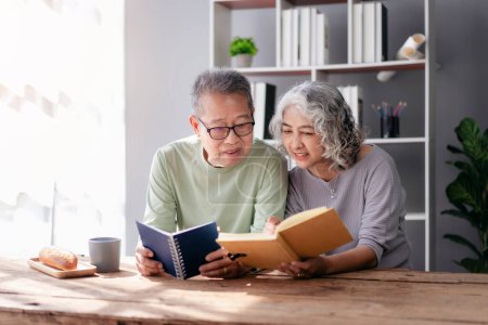 Photo for Senior couple embracing and reading a book together with enjoying in living room at home while spending. - Royalty Free Image