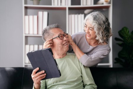 Photo for Senior woman putting glasses on beloved while sitting on the couch to watching movie on tablet and spending time to doing activity for relaxation in life retirement together at living room of home. - Royalty Free Image
