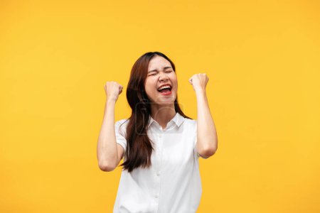 Téléchargez les photos : Jeune femme asiatique portant chemise blanche à manches courtes et levant les bras tout en souriant et en criant pour célébrer après avoir réussi à travailler isolé sur fond jaune. - en image libre de droit
