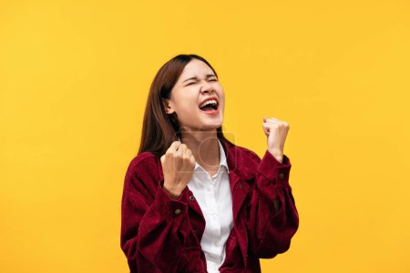 Téléchargez les photos : Jeune femme asiatique portant une veste rouge et grand sourire tout en serrant les poings et crié de joie après avoir réussi à travailler isolé sur fond jaune. - en image libre de droit