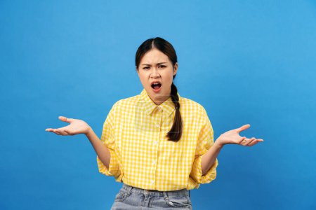 Foto de Joven mujer asiática vistiendo camisa amarilla con peinado trenzado y cara confusa mientras levanta las manos y grita a preguntar por qué aislado sobre fondo azul claro. - Imagen libre de derechos