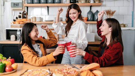 Foto de Grupo de amigos tomando un vaso de bebida y comiendo pizza italiana con pan juntos mientras disfrutan y levantan el brazo para celebrar en la fiesta en la cocina en casa. - Imagen libre de derechos