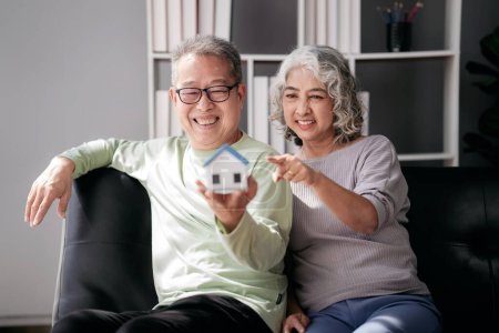 Photo for Senior couple holding house model for buying in life retirement while sitting on the couch and spending time to doing activity for relaxation together at living room of home. - Royalty Free Image