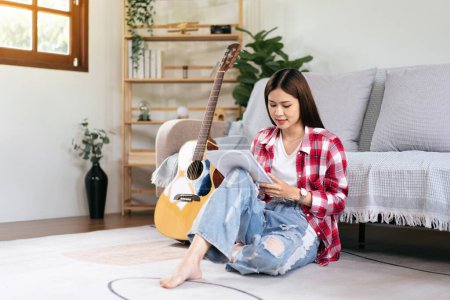 Photo for Young woman is compose the song and writing lyrics on notebook after playing guitar while sitting on the floor in living room at home. - Royalty Free Image