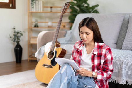 Photo for Young woman is compose the song and writing lyrics on notebook after playing guitar while sitting on the floor in living room at home. - Royalty Free Image