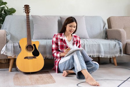 Téléchargez les photos : Jeune femme est de composer la chanson et d'écrire des paroles sur le carnet après avoir joué de la guitare tout en étant assis sur le sol dans le salon à la maison. - en image libre de droit