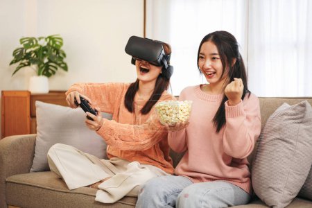 Téléchargez les photos : Jeune femme lesbienne couple portant pull et lunettes VR tout en tenant joystick pour jouer à un jeu vidéo avec l'expérience de la réalité virtuelle et manger du pop-corn pour passer du temps ensemble dans le salon. - en image libre de droit