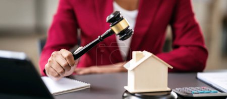 Photo for African american lawyer woman in suit holding judge hammer with house model and contract document on the desk for presentation about renting or buying and insurance housing in workspace modern. - Royalty Free Image
