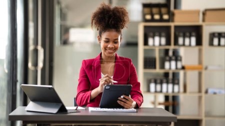 Téléchargez les photos : Femme d'affaires afro-américaine en costume en utilisant une tablette pour lire les informations comptables de nouvelles entreprises et de prendre des notes tout en travaillant et en réfléchissant à la stratégie de projet dans l'espace de travail moderne. - en image libre de droit