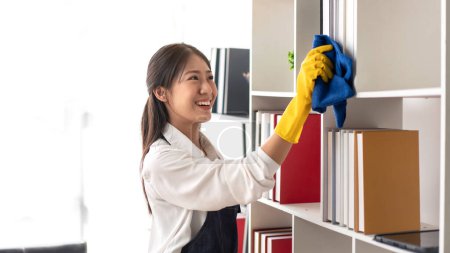 Téléchargez les photos : Femme au foyer dans le tablier portant des gants à l'aide de tissu en microfibre pour essuyer la bibliothèque et les meubles de nettoyage pendant le travail et le nettoyage dans la maison. - en image libre de droit