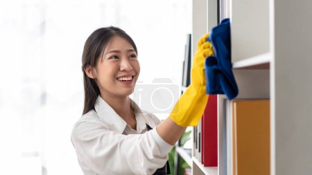 Téléchargez les photos : Femme au foyer dans le tablier portant des gants à l'aide de tissu en microfibre pour essuyer la bibliothèque et les meubles de nettoyage pendant le travail et le nettoyage dans la maison. - en image libre de droit