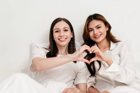 Photo for Young asian couple woman smiling and making heart sign gesture together while lying under blanket and leaning on comfortable pillows on the bed in bedroom at home. - Royalty Free Image
