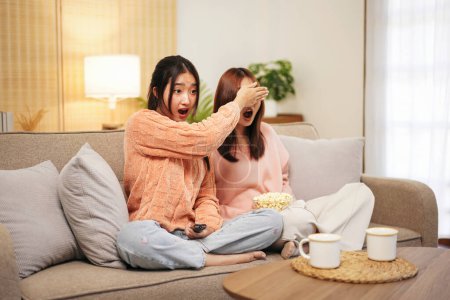 Photo for Young woman lesbian couple in sweater sits on comfortable sofa to covering eyes girlfriend and watching scary horror cinema with frightened face while spending time together in living room at home. - Royalty Free Image