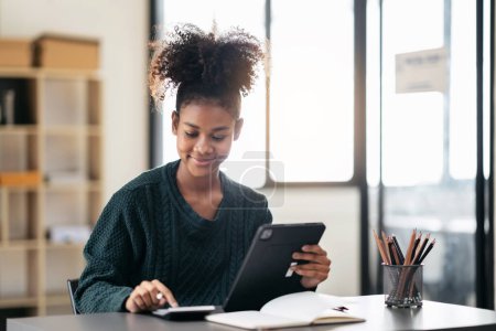 Téléchargez les photos : Femme étudiante afro-américaine en pull à l'aide d'une calculatrice pour faire des devoirs et écrire des informations sur une tablette numérique après avoir étudié la leçon en ligne tout en apprenant l'éducation à l'université. - en image libre de droit