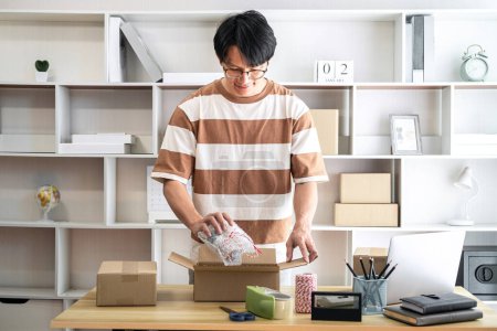 Téléchargez les photos : Homme entrepreneur emballe le produit dans des boîtes en carton pour se préparer à envoyer le client tout en travaillant la livraison d'achats en ligne au bureau à domicile. - en image libre de droit
