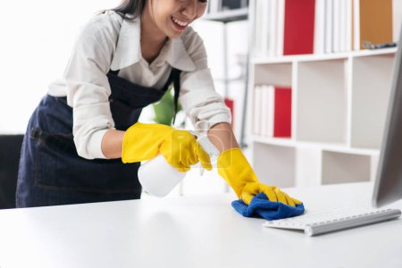 Photo for Housewife in apron wearing gloves to spraying hygiene spray on computer and using microfiber fabric to wiping cleanup the table while working and cleaning furniture in the house. - Royalty Free Image