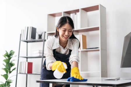 Photo for Housewife in apron wearing gloves to spraying hygiene spray on computer and using microfiber fabric to wiping cleanup the table while working and cleaning furniture in the house. - Royalty Free Image