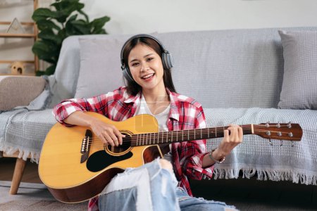 Téléchargez les photos : Jeune femme joue de la guitare et chante la chanson tout en portant un casque à écouter de la musique et assis sur le sol dans le salon à la maison. - en image libre de droit