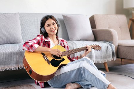 Photo for Young woman is playing guitar and singing the song while wearing headphone to listening music and sitting on the floor in living room at home. - Royalty Free Image