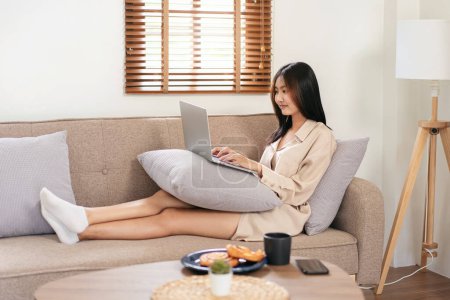 Téléchargez les photos : Femme entrepreneur est assis sur un grand canapé confortable et mettre un ordinateur portable sur l'oreiller pour taper des données commerciales tout en travaillant dans le salon à la maison. - en image libre de droit