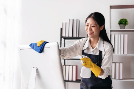 Téléchargez les photos : Femme au foyer dans le tablier portant des gants pour pulvériser pulvérisation d'hygiène sur l'ordinateur et en utilisant du tissu en microfibre pour essuyer les meubles de nettoyage pendant le travail et le nettoyage dans la maison. - en image libre de droit